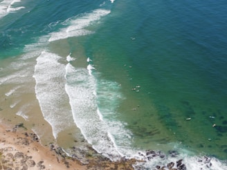 An aerial view of a coastline with waves breaking onto the shore. The turquoise and deep blue ocean transitions towards the sandy beach. Surfboards and surfers are visible in the water, waiting to catch waves.