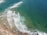 An aerial view of a coastline with waves breaking onto the shore. The turquoise and deep blue ocean transitions towards the sandy beach. Surfboards and surfers are visible in the water, waiting to catch waves.