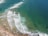 An aerial view of a coastline with waves breaking onto the shore. The turquoise and deep blue ocean transitions towards the sandy beach. Surfboards and surfers are visible in the water, waiting to catch waves.