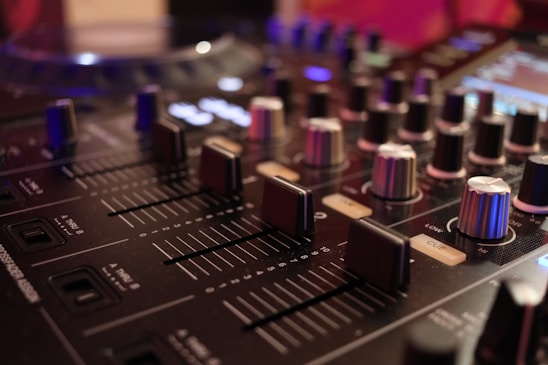 Close-up view of a professional DJ mixer with various knobs and sliders. The image captures the intricate details of the equipment, highlighting the texture of the knobs and the smooth surface of the sliders. Soft lighting adds a warm ambiance, casting gentle reflections on the mixer surface.