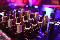 A close-up view of an audio mixing console with various knobs and sliders, set against a background of vivid, colorful lighting. The focus is on the numerous black and white dials used to adjust sound levels and frequencies.