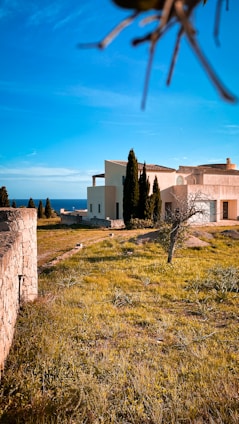 A modern beige house with flat roofs and multiple sections is surrounded by a grassy field with sparse greenery. Tall cypress trees line the property, and a stone wall runs along the left side leading toward the ocean visible in the background. The sky is clear blue, creating a serene and peaceful setting.