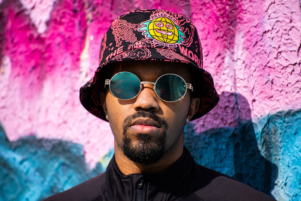a man wearing a hat and sunglasses in front of a colorful wall
