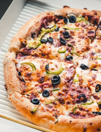 A close-up of a freshly baked pizza topped with melted cheese, black olives, green bell peppers, and slices of pepperoni. The crust is golden brown and slightly crispy, and the pizza is placed in an open cardboard delivery box.