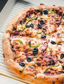 A close-up of a freshly baked pizza topped with melted cheese, black olives, green bell peppers, and slices of pepperoni. The crust is golden brown and slightly crispy, and the pizza is placed in an open cardboard delivery box.