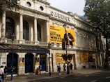 A grand historic theater building with intricate architectural details. The facade is adorned with large posters for the play 'Bitter Wheat' by David Mamet, starring John Malkovich. Pedestrians walk by on the sidewalk below, and trees cast shadows on the building.