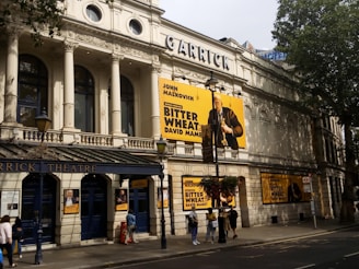A grand historic theater building with intricate architectural details. The facade is adorned with large posters for the play 'Bitter Wheat' by David Mamet, starring John Malkovich. Pedestrians walk by on the sidewalk below, and trees cast shadows on the building.