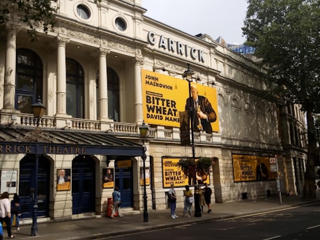 A grand historic theater building with intricate architectural details. The facade is adorned with large posters for the play 'Bitter Wheat' by David Mamet, starring John Malkovich. Pedestrians walk by on the sidewalk below, and trees cast shadows on the building.