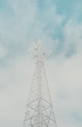A tall communication tower with lattice structure stands against a cloudy sky. The structure features multiple antennas and dishes, extending towards the sky. The atmosphere is serene with soft, pastel-colored clouds in the background.