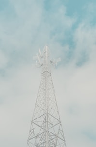 A tall communication tower with lattice structure stands against a cloudy sky. The structure features multiple antennas and dishes, extending towards the sky. The atmosphere is serene with soft, pastel-colored clouds in the background.