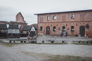 A rustic setting featuring an old brick building with a cobbled courtyard. Two metal knight statues stand against the wall of the building, which is adorned with small potted plants and old wagon wheels. To the left, a wooden structure with shields on display gives a medieval vibe. In the foreground, there is a campfire setup with benches surrounding it.