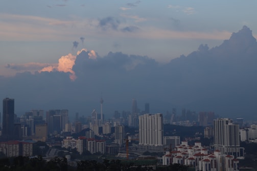 A city skyline at dusk with high-rise buildings and a prominent telecommunications tower. The sky is filled with dark clouds and patches of pinkish-orange light peeking through, creating a dramatic contrast.