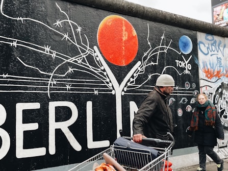 A colorful mural on a wall with the word 'BERLIN' prominently displayed alongside barbed wire imagery and a large red circular element, possibly representing a sun. Two people are walking in front of the wall, one of whom is pushing a shopping cart filled with goods.