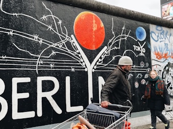 A colorful mural on a wall with the word 'BERLIN' prominently displayed alongside barbed wire imagery and a large red circular element, possibly representing a sun. Two people are walking in front of the wall, one of whom is pushing a shopping cart filled with goods.