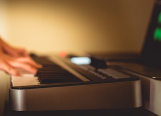 Hands are playing a keyboard or electric piano in a dimly lit room, suggesting a focus on music creation or practice. The background is softly blurred, and a screen with faint green lights is partially visible.