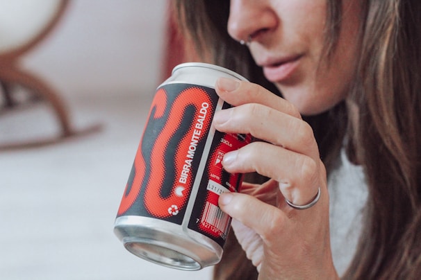 A person with long hair holds a can with a bold red and black design labeled 'Birra Monte Baldo'. The focus is on the hand and the can, while the person's face is partially visible. The atmosphere is casual and relaxed, with natural lighting.