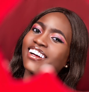 A person with a bright smile and glossy lips is captured in a close-up portrait. The background and surroundings are rich in red tones, complementing the makeup that includes pink eyeshadow.