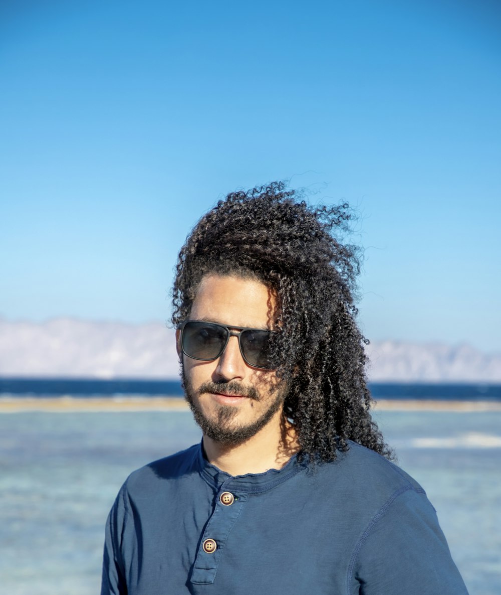 a man with long hair and sunglasses standing in front of a body of water
