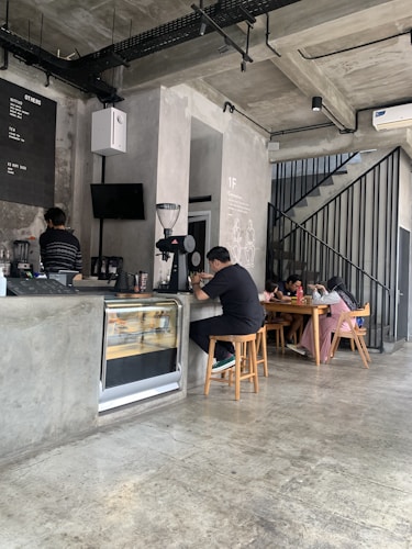 A modern cafe interior with concrete walls and floors, featuring an industrial design. A barista is standing behind the counter working on coffee equipment. There is a small display case integrated into the counter. A few patrons are sitting at a wooden table in the back, engaged in conversation.