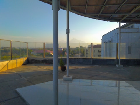 A covered rooftop area with glass railings offers a view of a distant mountain under a clear sky. The space is paved and has a structure with a metal roof. The morning light casts shadows on the tiles and highlights the simplicity and openness of the space.