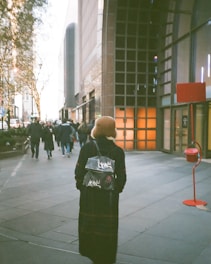 A person in a long coat and a hat walks down a city sidewalk. The individual carries a stylish backpack. Surrounding them are tall buildings with bright display windows. Holiday lights adorn the trees lining the street. Other pedestrians are visible in the background. A red donation kettle is placed on the right side of the sidewalk.