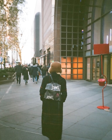 A person in a long coat and a hat walks down a city sidewalk. The individual carries a stylish backpack. Surrounding them are tall buildings with bright display windows. Holiday lights adorn the trees lining the street. Other pedestrians are visible in the background. A red donation kettle is placed on the right side of the sidewalk.