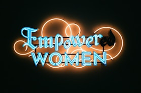 A neon sign with the words 'Empower Women' in bright blue letters against a dark background. The lettering is surrounded by an artistic design of neon orange loops, adding a decorative touch to the sign.