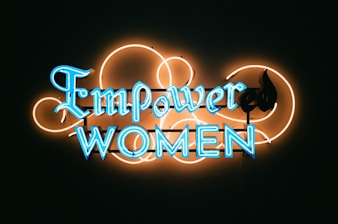 A neon sign with the words 'Empower Women' in bright blue letters against a dark background. The lettering is surrounded by an artistic design of neon orange loops, adding a decorative touch to the sign.