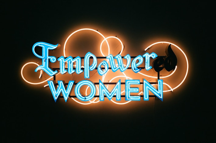 A neon sign with the words 'Empower Women' in bright blue letters against a dark background. The lettering is surrounded by an artistic design of neon orange loops, adding a decorative touch to the sign.