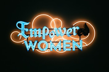 A neon sign with the words 'Empower Women' in bright blue letters against a dark background. The lettering is surrounded by an artistic design of neon orange loops, adding a decorative touch to the sign.
