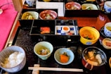 A variety of small, beautifully presented dishes are arranged on a black tray, featuring an assortment of Japanese cuisine. There are bowls with rice, fish, soup, pickled vegetables, and a half-boiled egg. The food is served in elegant, colorful dishes, including blue, green, and red tones. Chopsticks are placed neatly on a rest in front of the tray.