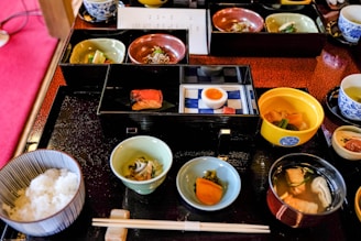 A variety of small, beautifully presented dishes are arranged on a black tray, featuring an assortment of Japanese cuisine. There are bowls with rice, fish, soup, pickled vegetables, and a half-boiled egg. The food is served in elegant, colorful dishes, including blue, green, and red tones. Chopsticks are placed neatly on a rest in front of the tray.
