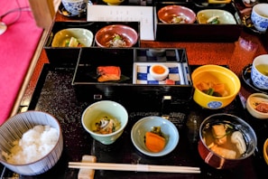 A variety of small, beautifully presented dishes are arranged on a black tray, featuring an assortment of Japanese cuisine. There are bowls with rice, fish, soup, pickled vegetables, and a half-boiled egg. The food is served in elegant, colorful dishes, including blue, green, and red tones. Chopsticks are placed neatly on a rest in front of the tray.