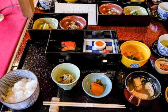 A variety of small, beautifully presented dishes are arranged on a black tray, featuring an assortment of Japanese cuisine. There are bowls with rice, fish, soup, pickled vegetables, and a half-boiled egg. The food is served in elegant, colorful dishes, including blue, green, and red tones. Chopsticks are placed neatly on a rest in front of the tray.
