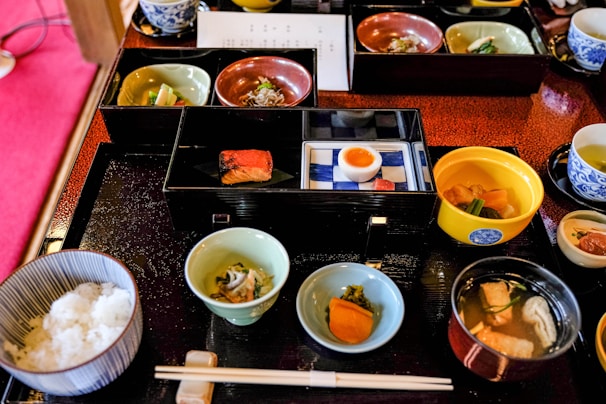 A variety of small, beautifully presented dishes are arranged on a black tray, featuring an assortment of Japanese cuisine. There are bowls with rice, fish, soup, pickled vegetables, and a half-boiled egg. The food is served in elegant, colorful dishes, including blue, green, and red tones. Chopsticks are placed neatly on a rest in front of the tray.