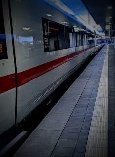 A modern high-speed train is stationed at a platform, with a sleek white exterior and red stripe running along its side. The train station appears quiet, with a tiled platform and signs visible in the background.