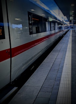 A modern high-speed train is stationed at a platform, with a sleek white exterior and red stripe running along its side. The train station appears quiet, with a tiled platform and signs visible in the background.