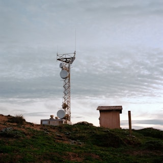 A telecommunications tower stands on a grassy hilltop, surrounded by a few small buildings. The sky is overcast with a layer of clouds, creating a calm and serene atmosphere.
