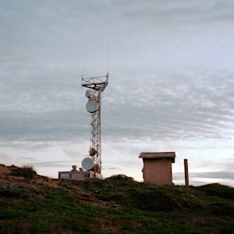 A telecommunications tower stands on a grassy hilltop, surrounded by a few small buildings. The sky is overcast with a layer of clouds, creating a calm and serene atmosphere.