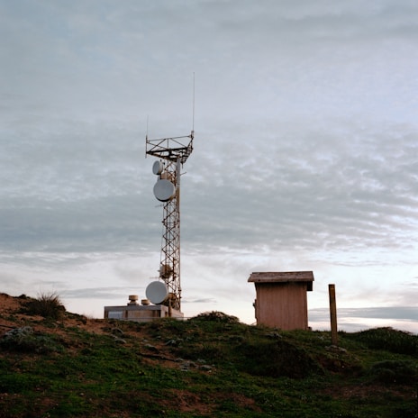 A telecommunications tower stands on a grassy hilltop, surrounded by a few small buildings. The sky is overcast with a layer of clouds, creating a calm and serene atmosphere.
