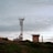 A telecommunications tower stands on a grassy hilltop, surrounded by a few small buildings. The sky is overcast with a layer of clouds, creating a calm and serene atmosphere.