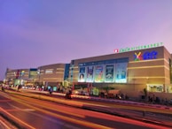 An illuminated shopping mall at dusk with large signages and advertisements on the facade. The sky is a serene shade of purple, and vibrant streaks of light trail on the road in front, likely from passing vehicles. The building features a prominent hypermarket sign at the top and well-lit storefronts.