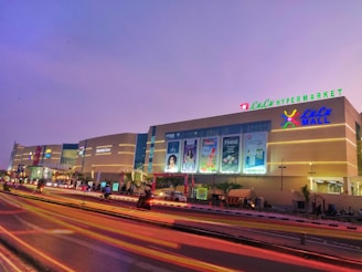 An illuminated shopping mall at dusk with large signages and advertisements on the facade. The sky is a serene shade of purple, and vibrant streaks of light trail on the road in front, likely from passing vehicles. The building features a prominent hypermarket sign at the top and well-lit storefronts.