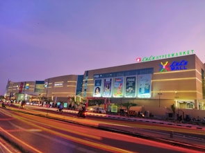 An illuminated shopping mall at dusk with large signages and advertisements on the facade. The sky is a serene shade of purple, and vibrant streaks of light trail on the road in front, likely from passing vehicles. The building features a prominent hypermarket sign at the top and well-lit storefronts.
