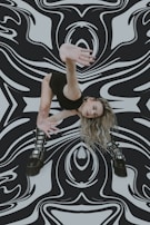 A woman in a dynamic, dance-like pose with her arm extended towards the camera. She has long, wavy blonde hair and wears a form-fitting black outfit and chunky black boots. The background features a bold, black-and-white abstract pattern that appears to swirl and warp around her.
