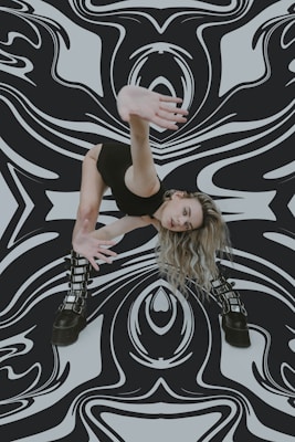 A woman in a dynamic, dance-like pose with her arm extended towards the camera. She has long, wavy blonde hair and wears a form-fitting black outfit and chunky black boots. The background features a bold, black-and-white abstract pattern that appears to swirl and warp around her.