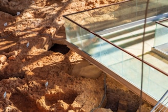 An archaeological site with excavated earth showing numbered markers in various pits. A modern glass walkway is built above the excavation, allowing visitors to view the site without disturbing it. The ground is uneven and has a reddish-brown color, with shadows cast on the surface.