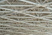 A complex network of interconnected beams and girders made of metal, arranged in a geometric pattern. The structure is intricate, with multiple layers and angles lending a sense of industrial and architectural design.