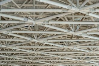 A complex network of interconnected beams and girders made of metal, arranged in a geometric pattern. The structure is intricate, with multiple layers and angles lending a sense of industrial and architectural design.