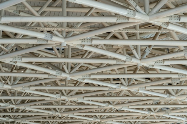 A complex network of interconnected beams and girders made of metal, arranged in a geometric pattern. The structure is intricate, with multiple layers and angles lending a sense of industrial and architectural design.